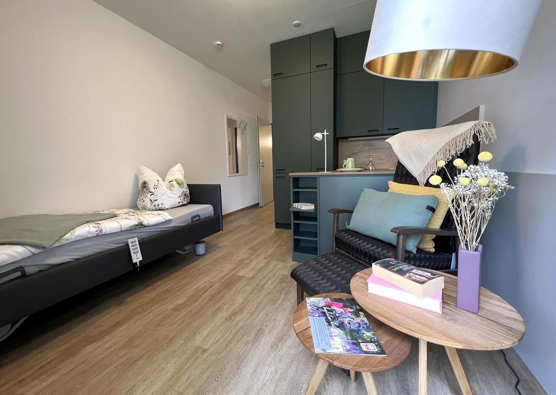 Zimmer mit Bett auf der linken Seite, Küchenzeile (rechts) mit dunkelgrünen Fronten und davor einem dunklen Sessel mit Beistelltisch in Holzoptik und einer gold-weißen Lampe.