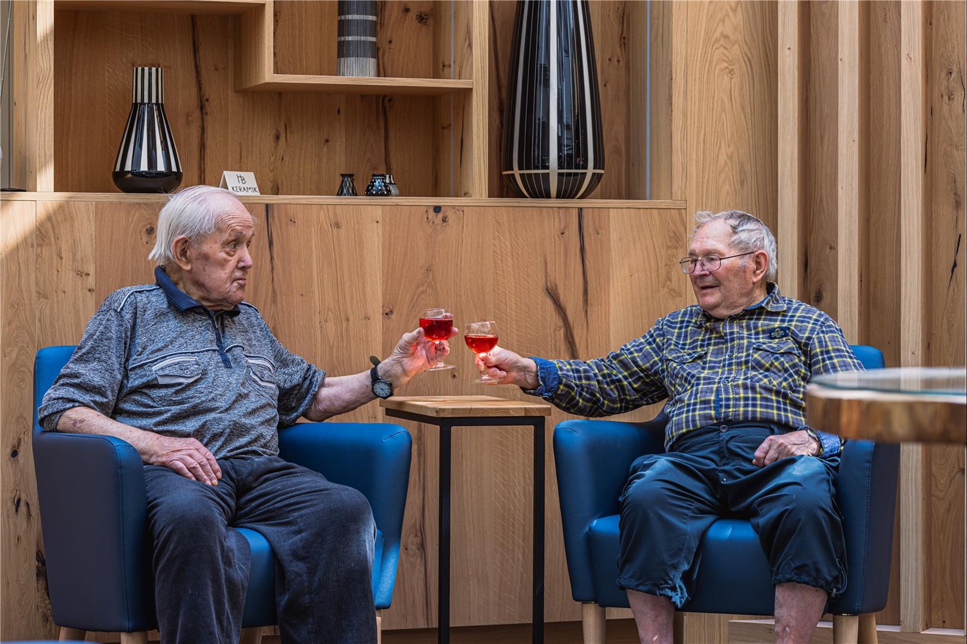 Zwei Männer sitzen im Foyer in Velten vor der neu gestalteten Rückwand und stoßen an.