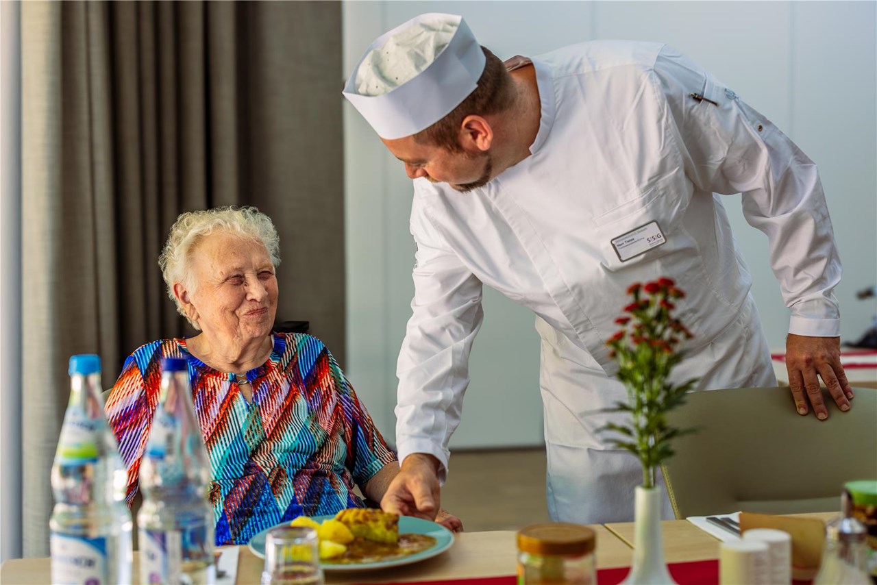 Koch (rechts) reicht Seniorin, die am Tisch sitzt einen Teller mit frisch zubereitetem Essen.