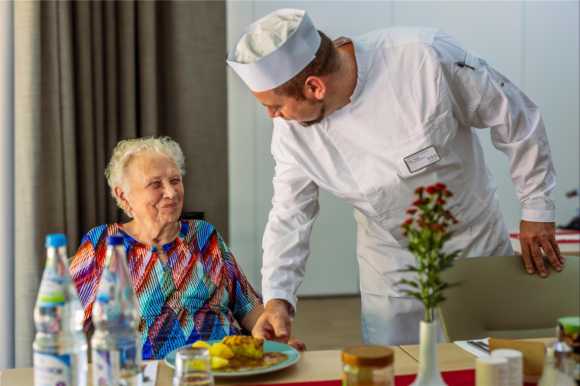Koch (rechts) reicht Seniorin, die am Tisch sitzt einen Teller mit frisch zubereitetem Essen.